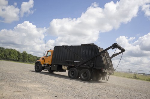 A large yellow and black rubbish collection truck with a metal hopper and tipping mechanism is parked on a gravel driveway under a partly cloudy sky. The truck is positioned to the left of the image, with the rear hopper slightly raised and tilted, suggesting it is in the process of unloading waste materials. The vehicle's body is predominantly yellow, with black for the hopper and tipping platform, and appears to be in good condition. The gravel surface surrounds the truck, with a flat open field extending into the distance, and a line of trees visible on the horizon. The scene is outdoors during daylight, with natural light illuminating the scene clearly. This image illustrates waste removal services, such as those offered by Business Waste Removal Golders Green, in a rural or suburban environment typical of the London boroughs, and emphasizes the practical aspect of rubbish collection and disposal on-site.